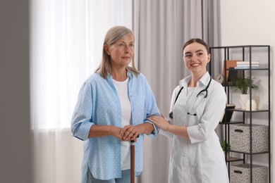 Young healthcare worker assisting senior woman with walking cane indoors