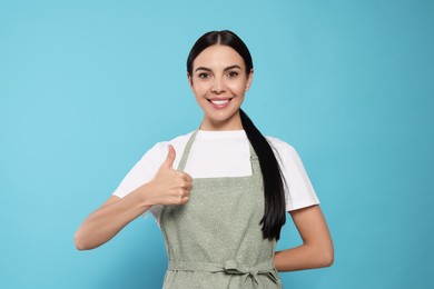 Young woman in grey apron showing thumb up on light blue background,