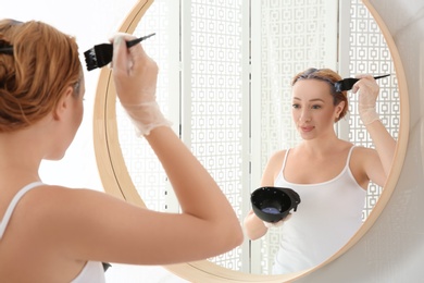 Photo of Young woman applying hair dye on roots in front of mirror at home