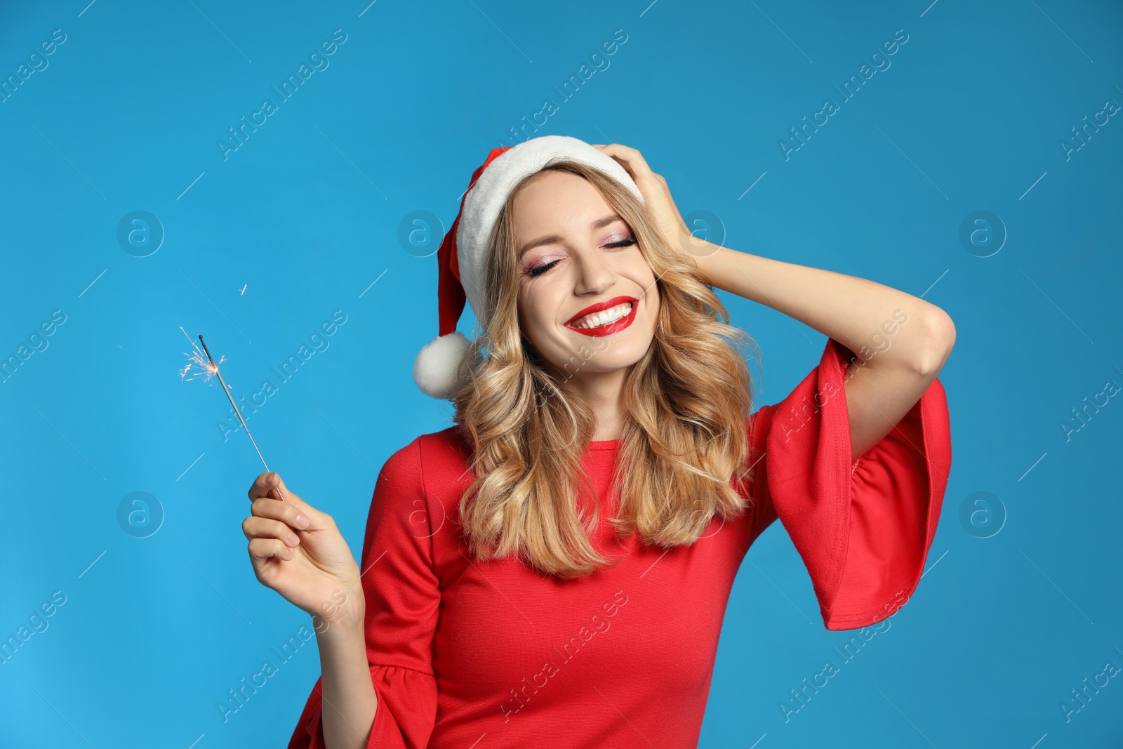 Photo of Happy young woman wearing Santa hat with sparkler on light blue background. Christmas celebration