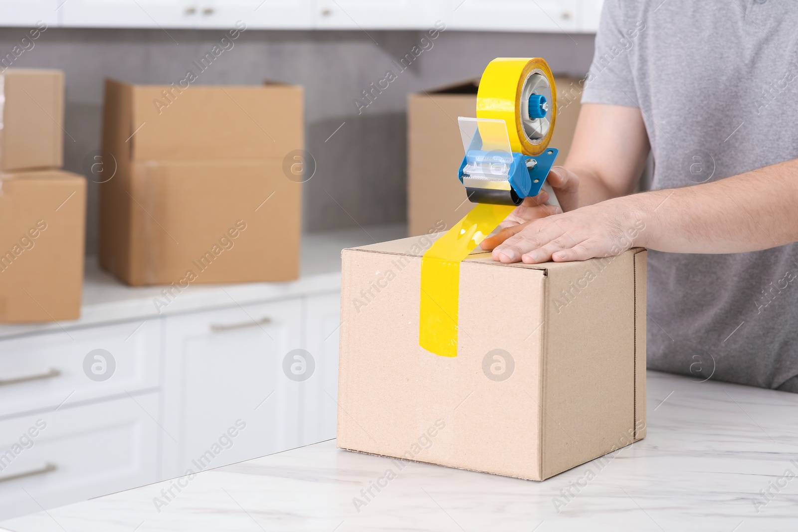 Photo of Man taping box with adhesive tape dispenser in kitchen, closeup