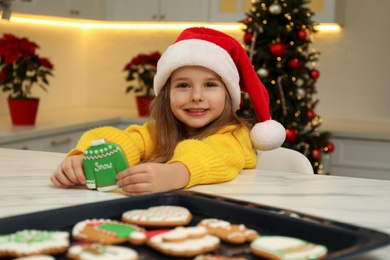 Photo of Cute little girl with freshly baked Christmas gingerbread cookie at table indoors