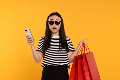 Surprised woman with shopping bags and smartphone on yellow background