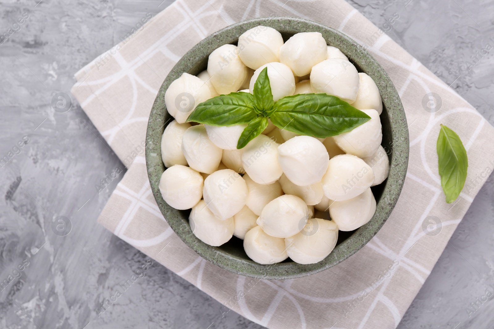 Photo of Tasty mozzarella balls and basil leaves in bowl on grey table, top view. Space for text