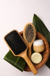 Wicker basket with wooden hairbrushes, cosmetic products and green leaves on white background, top view