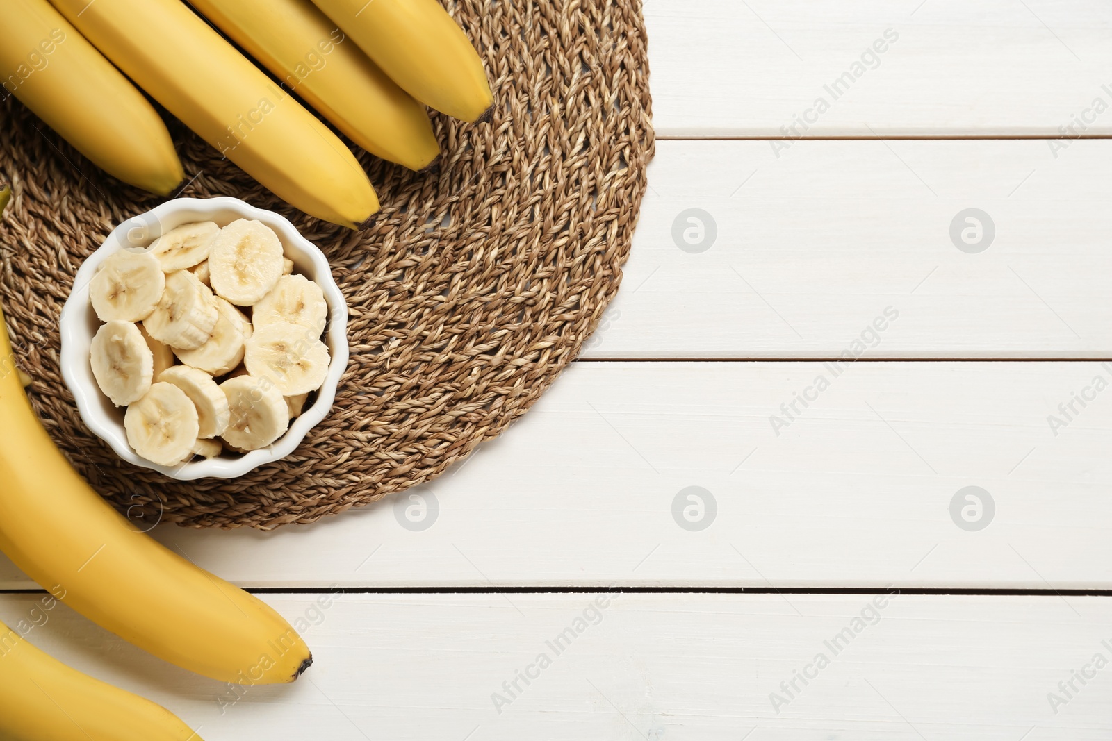 Photo of Bowl with cut bananas near whole fruits on white wooden table, flat lay. Space for text