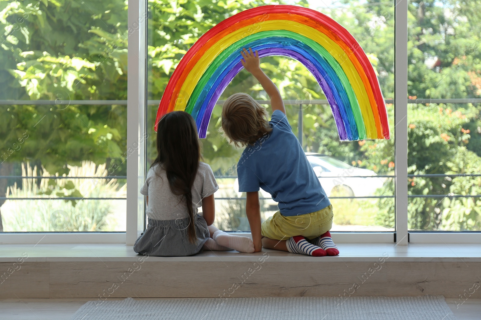 Photo of Little children sitting near rainbow painting on window indoors. Stay at home concept