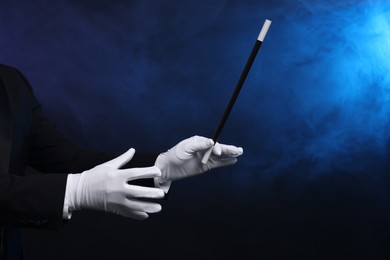 Magician holding wand in smoke on dark background, closeup. Space for text