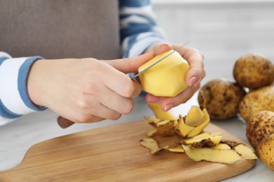 Woman peeling fresh potato with knife at white table, closeup