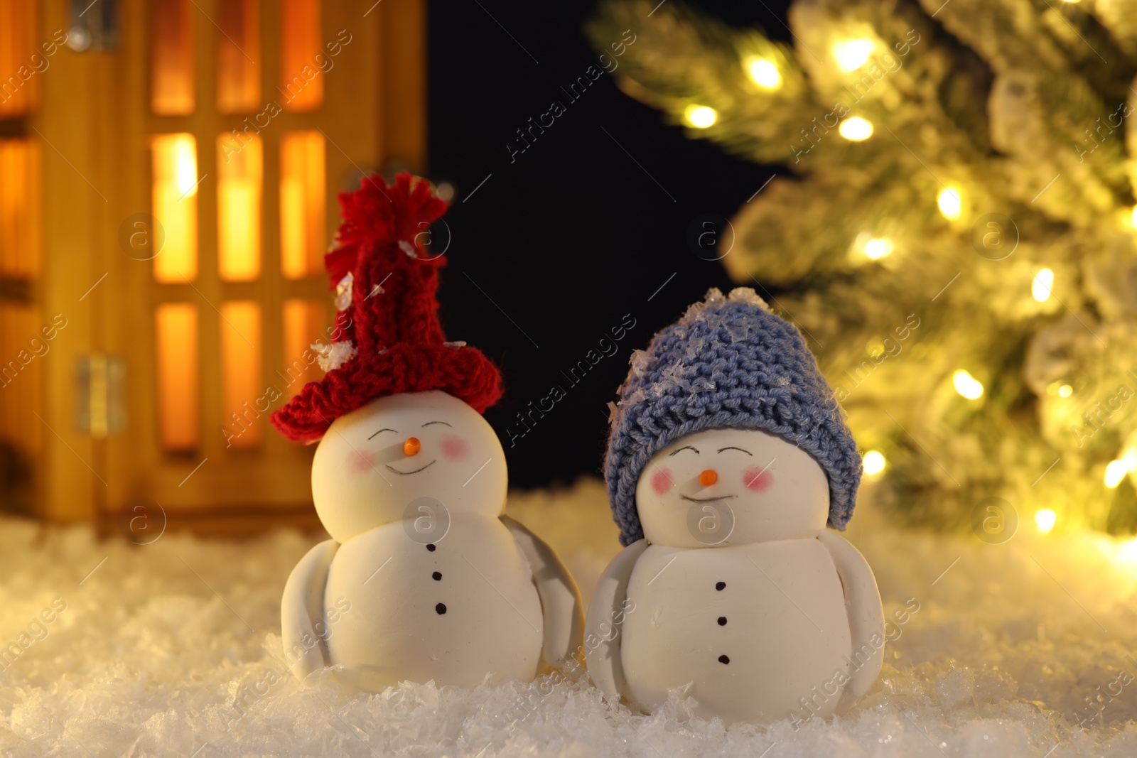 Photo of Cute decorative snowmen, lantern and Christmas tree on artificial snow against dark background