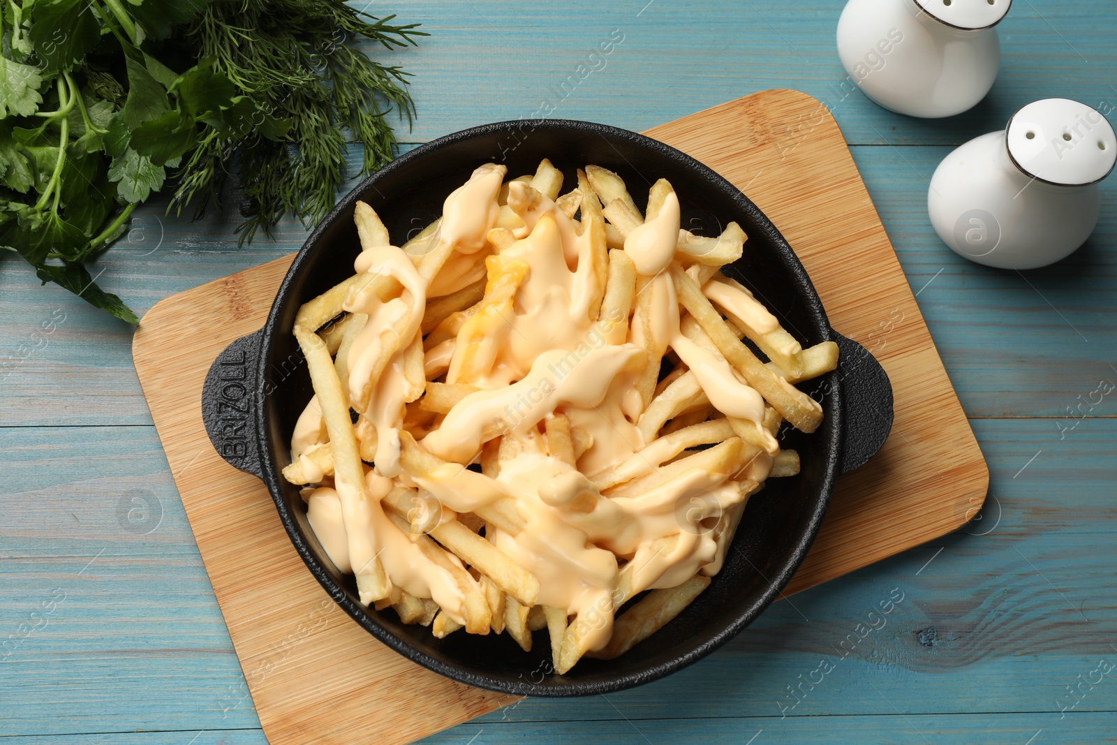 Photo of Delicious french fries with cheese sauce in bowl, greens and spice shakers on light blue wooden table, flat lay