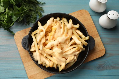 Photo of Delicious french fries with cheese sauce in bowl, greens and spice shakers on light blue wooden table, flat lay