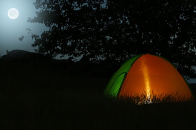 Photo of Modern tent lit from inside in wilderness at night, space for text. Overnight camping