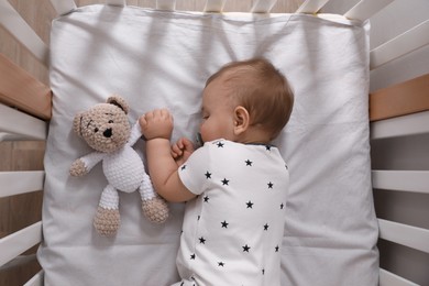 Photo of Adorable little baby with pacifier and toy sleeping in crib indoors, top view