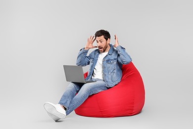 Emotional man with laptop sitting on beanbag chair against light grey background