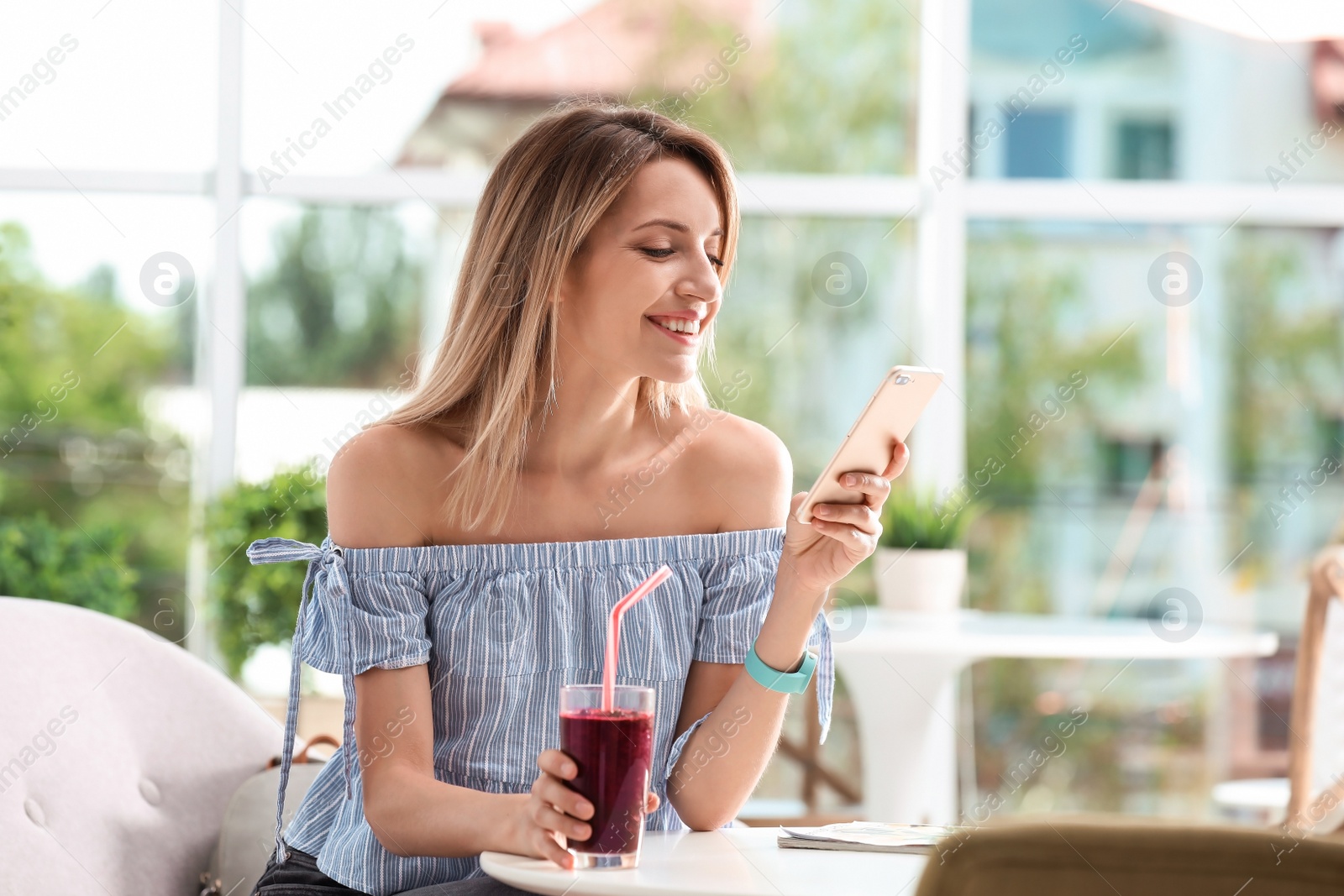 Photo of Young woman using mobile phone while drinking tasty healthy smoothie at table, indoors