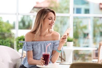 Young woman using mobile phone while drinking tasty healthy smoothie at table, indoors