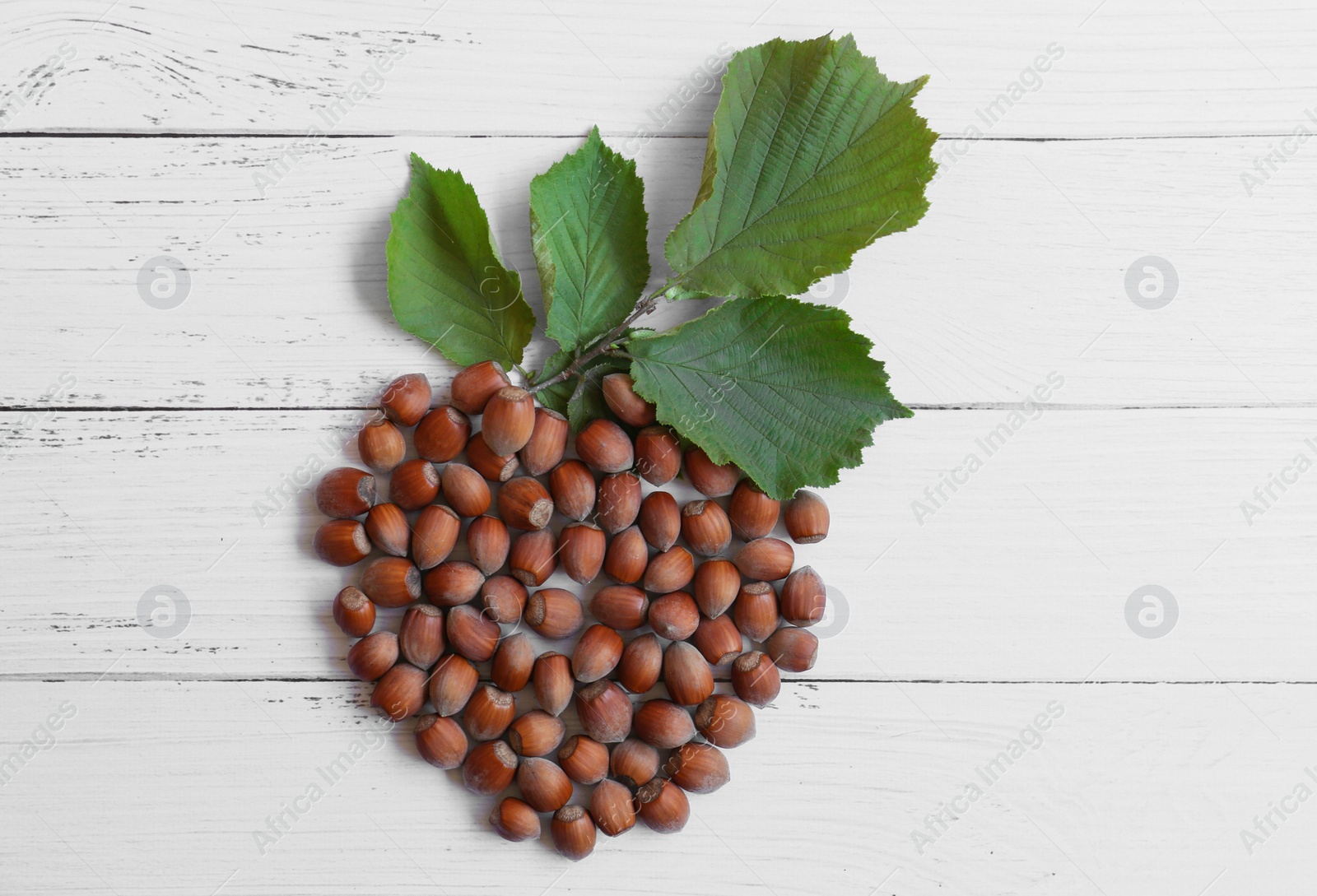 Photo of Hazelnuts and branch with leaves in shape of nut on white wooden table, flat lay