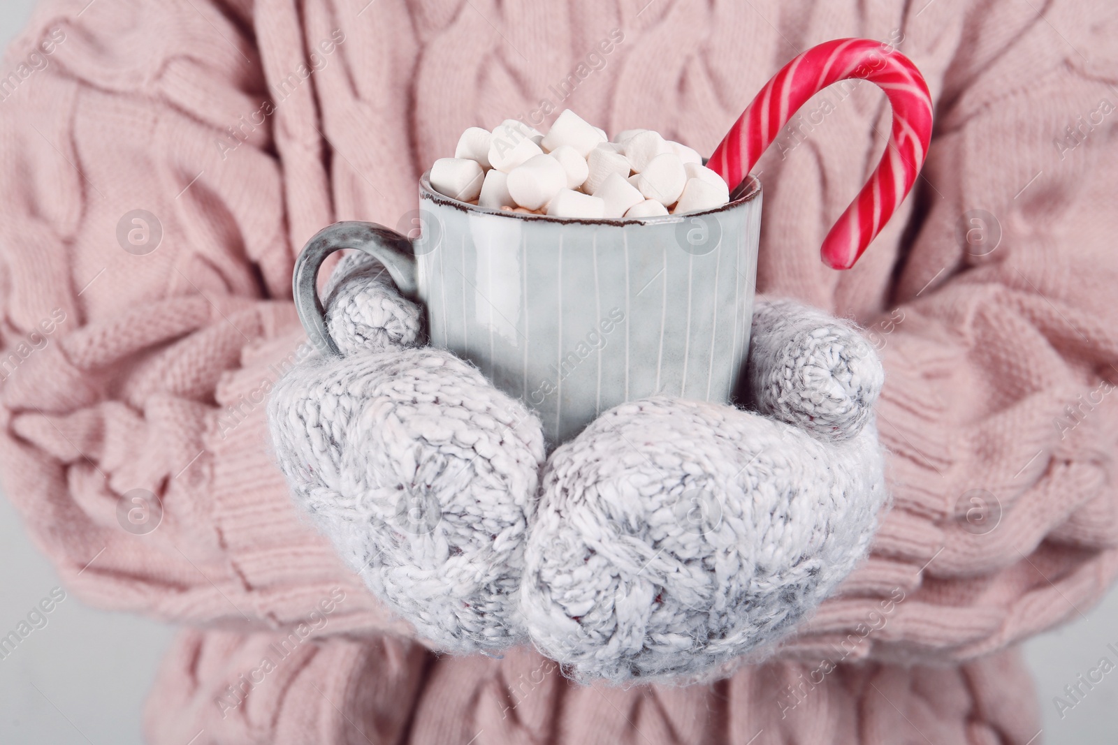 Photo of Woman in knitted mittens holding cup of delicious hot chocolate with marshmallows and candy cane, closeup