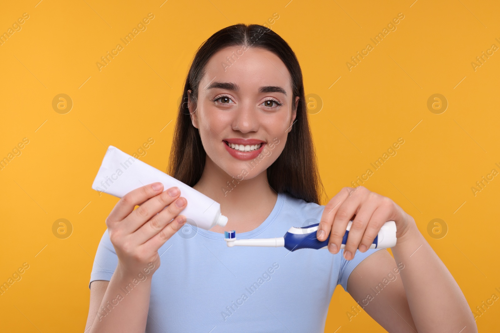 Photo of Happy young woman squeezing toothpaste from tube onto electric toothbrush on yellow background
