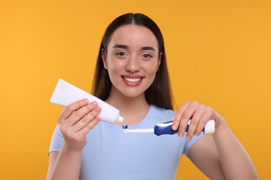 Happy young woman squeezing toothpaste from tube onto electric toothbrush on yellow background