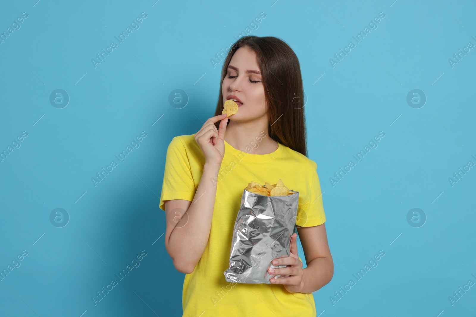 Photo of Pretty young woman eating tasty potato chips on light blue background