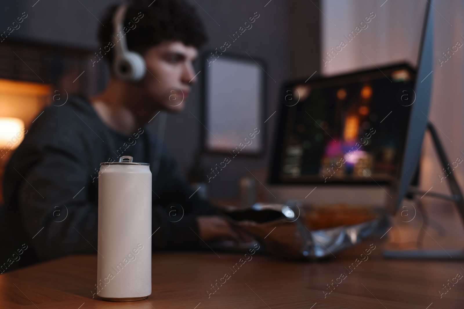 Photo of Young man with energy drink playing video game at wooden desk indoors, focus on can