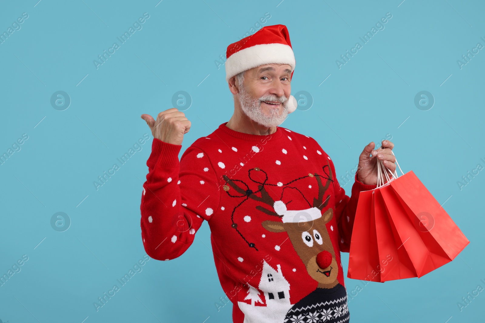 Photo of Happy senior man in Christmas sweater and Santa hat with shopping bags on light blue background