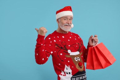 Photo of Happy senior man in Christmas sweater and Santa hat with shopping bags on light blue background