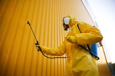 Photo of Person in hazmat suit disinfecting building wall with sprayer outdoors. Surface treatment during coronavirus pandemic