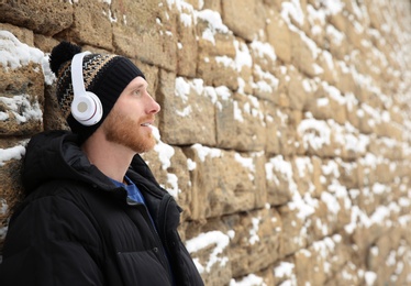 Photo of Young man listening to music with headphones near stone wall. Space for text