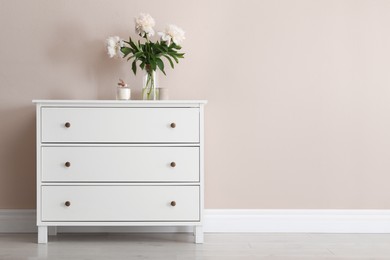 Photo of White chest of drawers with bouquet and candle near beige wall. Space for text