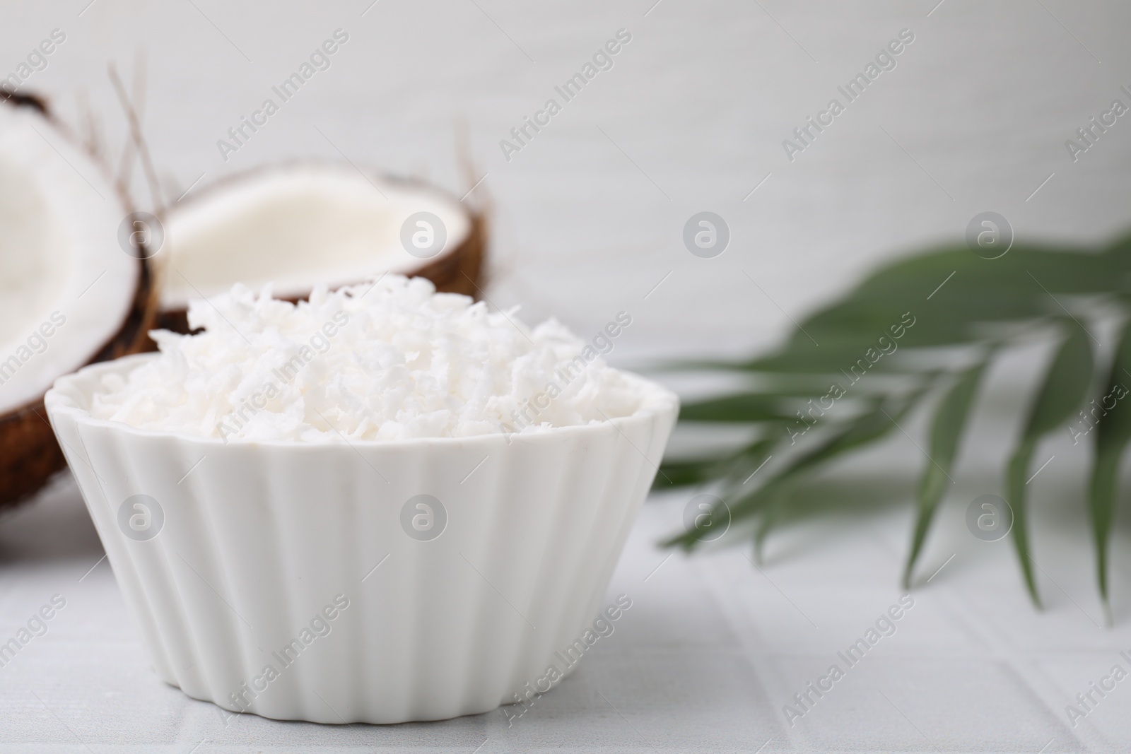 Photo of Coconut flakes in bowl, nuts and palm leaf on white tiled table, closeup