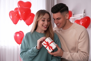 Man presenting gift to his girlfriend in room decorated with heart shaped balloons. Valentine's day celebration