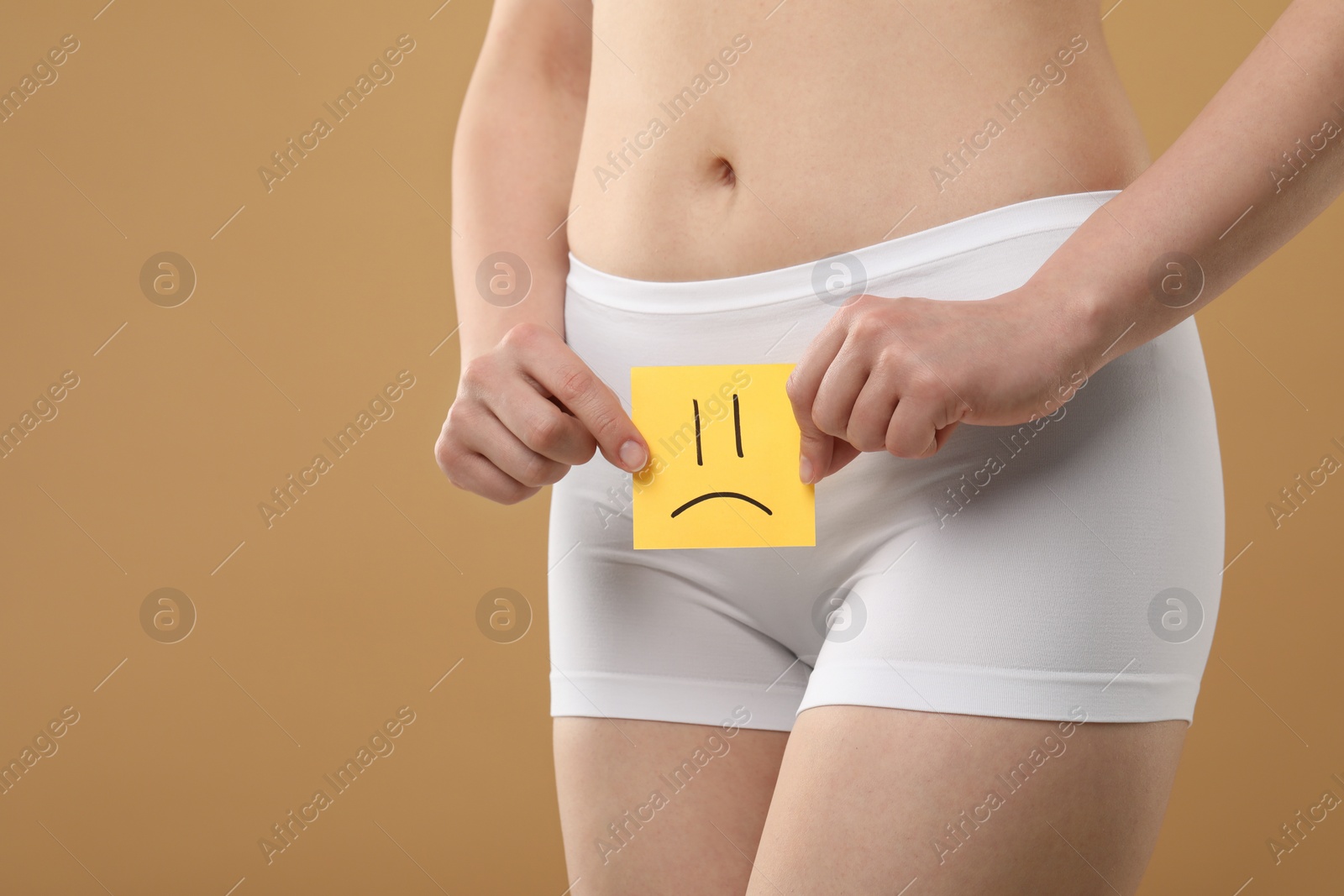 Photo of Cystitis. Woman holding sticky note with drawn sad face on beige background, closeup. Space for text