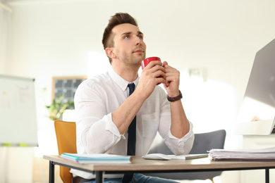 Young businessman with cup of drink relaxing at table in office during break