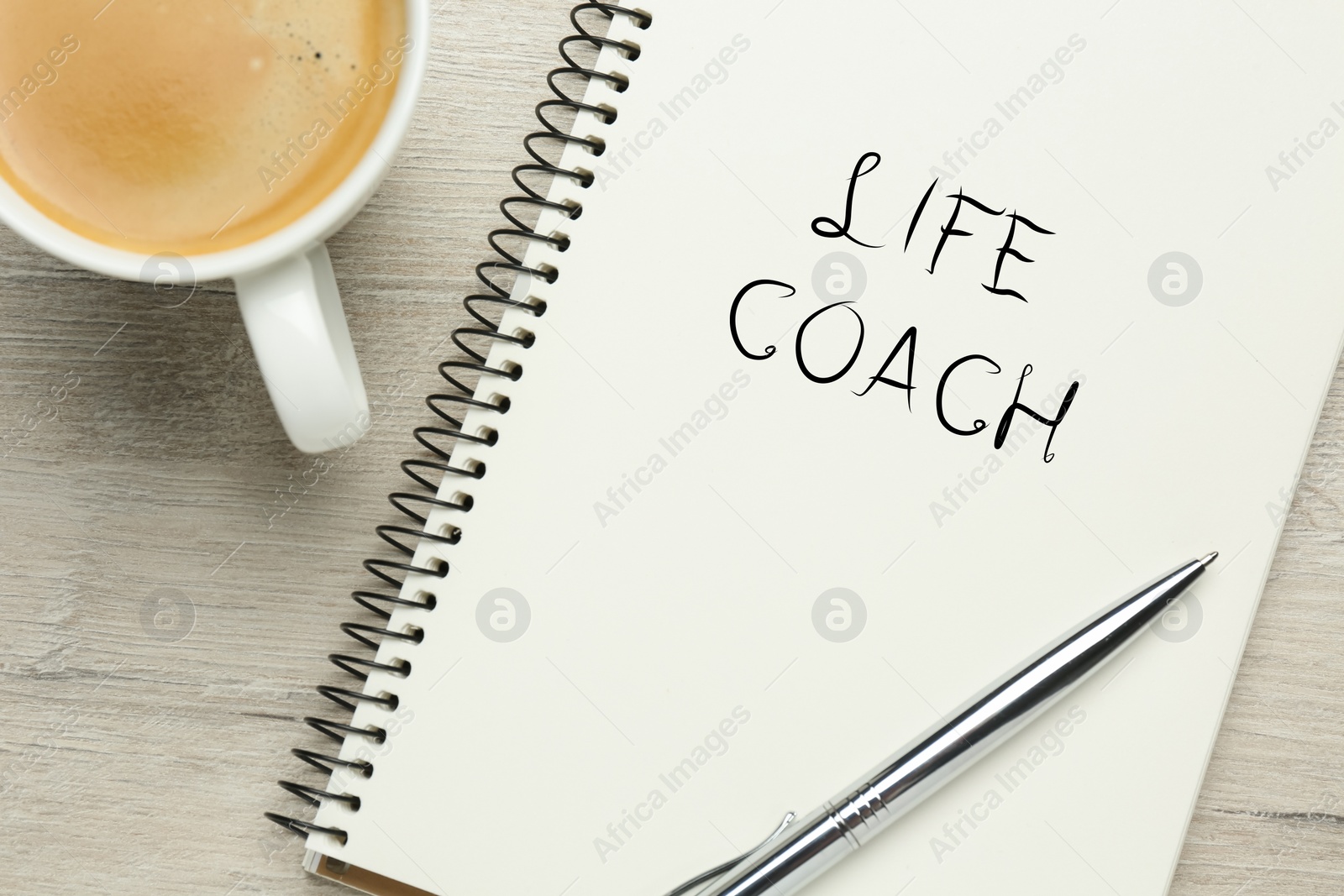 Image of Phrase Life Coach written in notebook, pen and cup coffee on white wooden table, flat lay