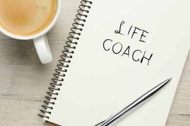 Image of Phrase Life Coach written in notebook, pen and cup coffee on white wooden table, flat lay