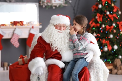 Photo of Little girl whispering in authentic Santa Claus' ear indoors