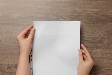 Woman putting paper sheet into punched pocket at wooden table, top view