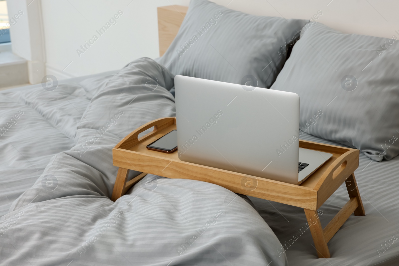 Photo of Wooden tray table with laptop and smartphone on bed indoors