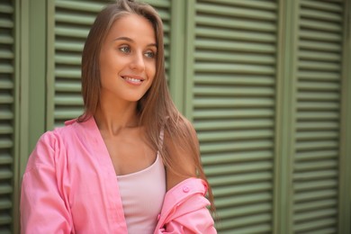 Photo of Beautiful young woman in stylish pink shirt near shutters, space for text