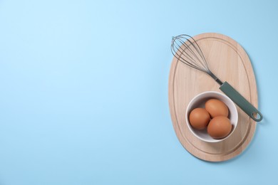 Photo of Metal whisk and raw eggs on light blue background, top view. Space for text