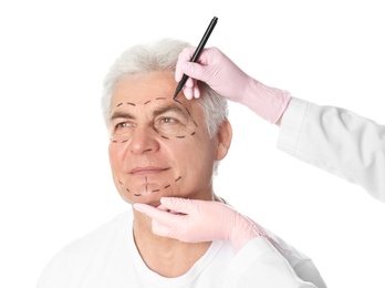 Doctor drawing marks on mature man's face for cosmetic surgery operation against white background