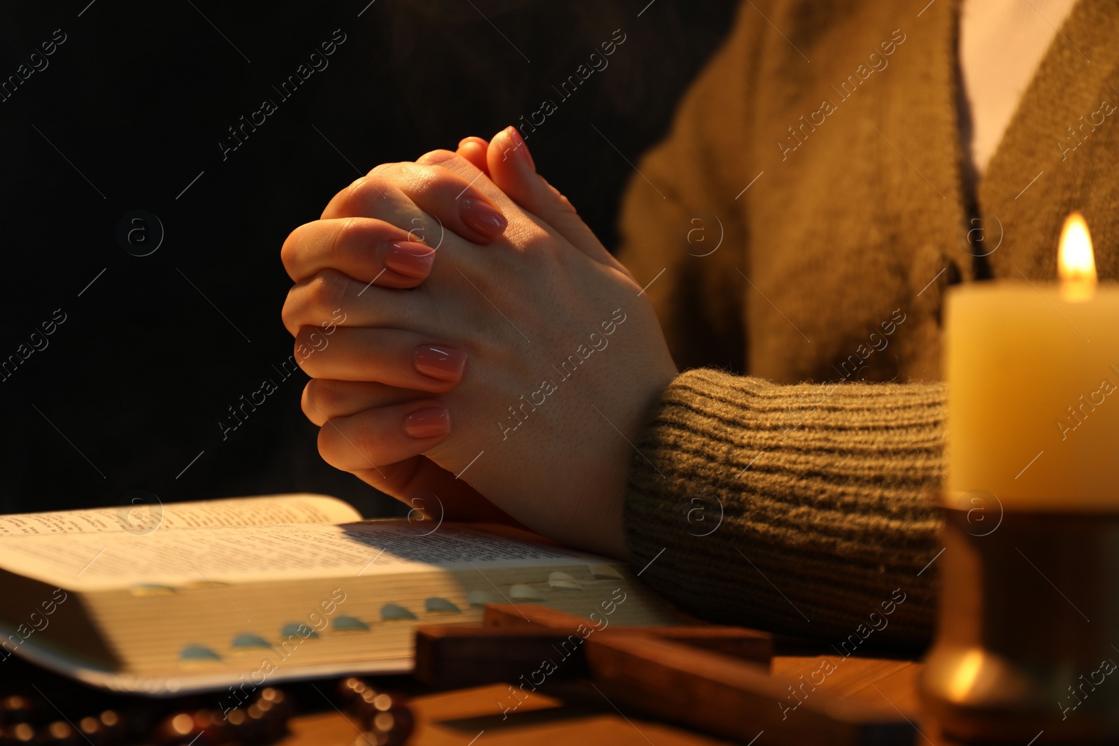 Photo of Woman praying at table with burning candle and Bible, closeup