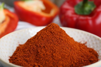 Bowl with aromatic paprika powder near fresh bell peppers, closeup