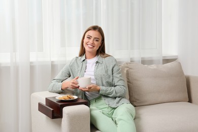 Photo of Happy woman holding cup of drink on sofa armrest wooden table at home
