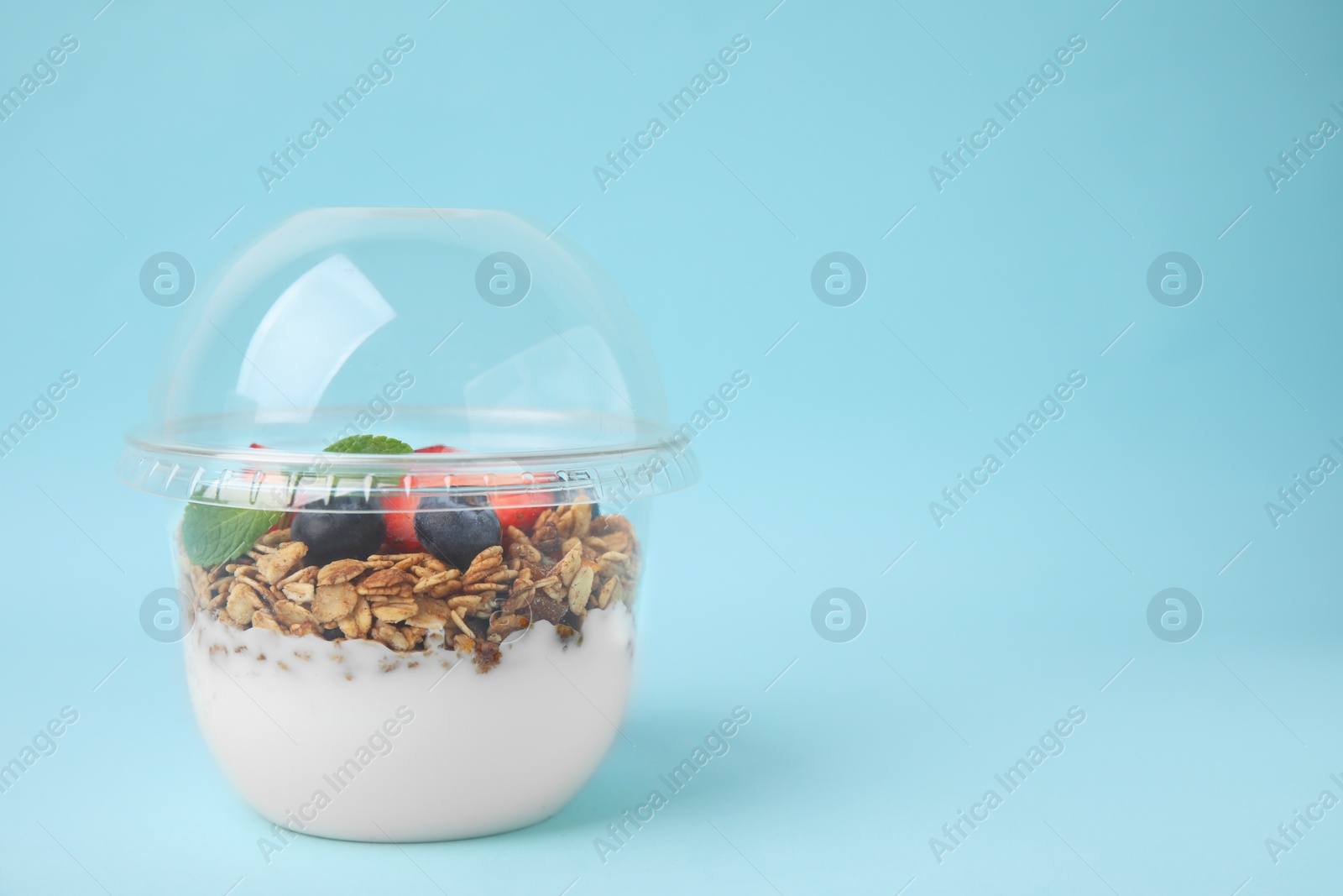 Photo of Tasty granola with berries and yogurt in plastic cup on light blue background, space for text