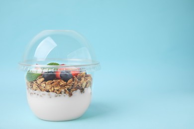 Photo of Tasty granola with berries and yogurt in plastic cup on light blue background, space for text