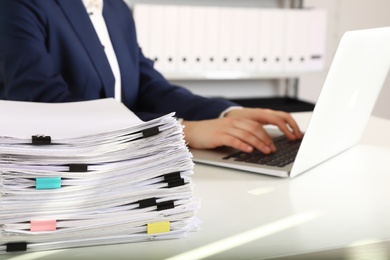 Stack of documents and woman working with laptop at table in office, closeup